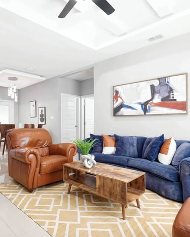 Open-plan living room with blue sofa, cognac leather chair, wooden coffee table, and kitchen with white cabinets and black bar stools