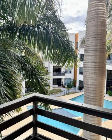 Modern apartment complex courtyard with two turquoise swimming pools, multi-story white buildings with black balconies, and palm tree.