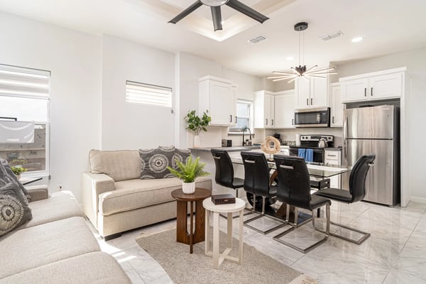 Open-plan living room and kitchen with beige sofa, dining table, stainless steel appliances, and white cabinetry