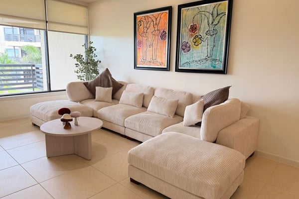 Modern living room with cream sectional sofa, round white coffee table, framed botanical artwork, and glass doors opening to balcony.